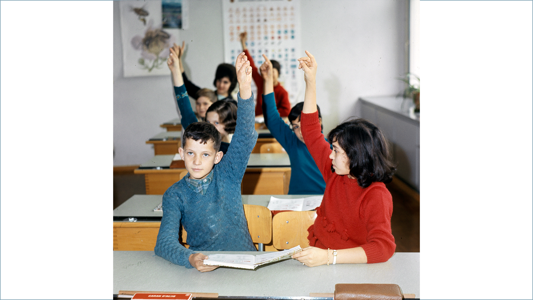 Kinder sitzen in der Schulstube an ihren Tischen und heben die Hand