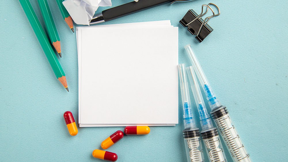 Image symbolique: vue d'en haut sur une feuille de papier, autour de laquelle se trouvent pilule, seringues et crayons